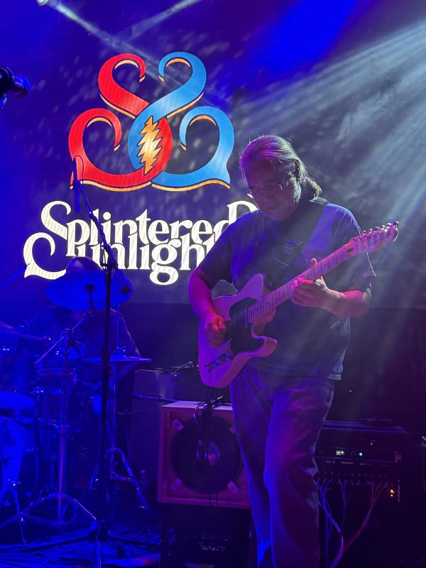 Member of Splintered Sunlight playing electric guitar in front of their logo.